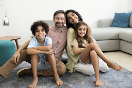 Happy Latin couple of parents and two cheerful kids sitting on carpeted floor at cozy home, looking at camera, posing for family portrait, smiling, laughing, enjoying childhood, parenthoodの写真素材