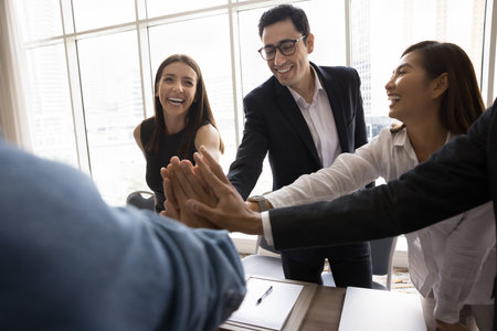 Excited positive diverse business partners celebrating successful collaboration, startup. Office friends joining hands in high five over meeting table, laughing, promoting teamwork, partnershipの写真素材