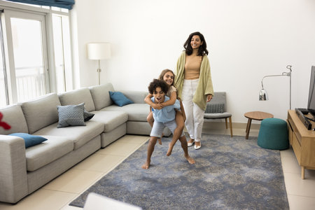 Cheerful active sibling children and Latin mom playing active games at home, dancing to music in living room, singing songs, shouting. Excited strong brother piggybacking sisterの写真素材