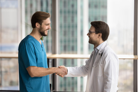 Happy friendly practitioner and surgeon doctors men in uniforms shaking hands, thanking for cooperation, good job, celebrating teamwork success. Medical colleagues giving handshakesの写真素材