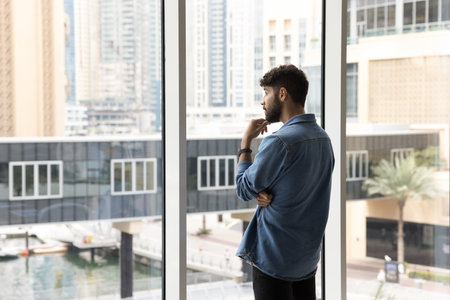 Thoughtful serious handsome young Arab man in casual informal clothes standing at large window,...