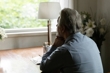 Far away in thoughts. Rear view of aged hoary male sit by table at home alone look at window ponder...