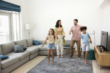 Cheerful middle aged parents and two preteen kids dancing in cozy flat interior, enjoying funny leisure, movement, family activity, hopping to music, singing, laughing, Full length shotの写真素材