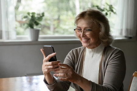 Aged smartphone user. Happy positive senior female pensioner holding mobile phone enjoy using web app reading news texting message answering call. Confident retired lady checking email chatting onlineの写真素材