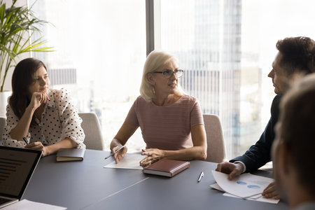 Confident elder businesswoman talking to younger business partners on negotiation meeting,...