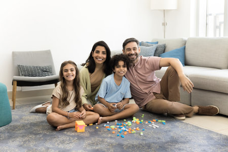Cheerful parents and two happy children sitting on warm floor at toy construction blocks, looking at...