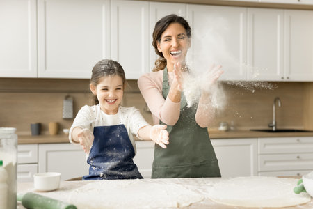 Excited mom and cheerful little daughter girl in aprons throwing flour while baking in home kitchen,...