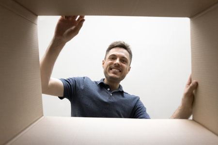 Positive young man opening large cardboard shipping box, looking inside, smiling, unpacking parcel...