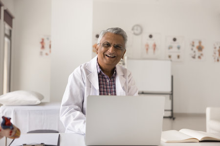 Happy mature senior Indian doctor man wearing white uniform and glasses, sitting at clinic workplace table, working at laptop, looking at camera, laughing, smiling, posing for portraitの写真素材