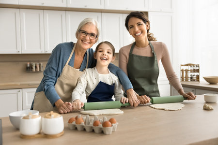 Family portrait of happy beautiful granny, mom and cheerful cute child girl in aprons preparing pastry, rolling dough on kitchen table for baking dessert, looking at camera, smilingの写真素材
