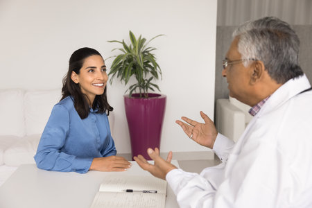 Happy young woman visiting senior Indian doctor office, consulting practitioner about health, healthcare service, smiling. Elder physician man talking to female Latin patient at workplace tableの写真素材