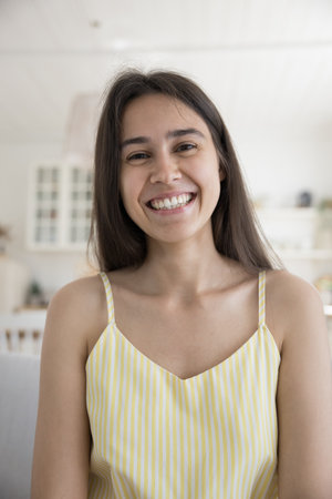 Happy young female look at camera sit on sofa at home make video conference to family living abroad,...