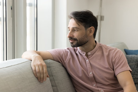 Positive thoughtful relaxed middle aged man sitting on comfortable couch, looking at window away,...