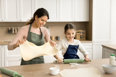 Happy sweet kid girl helping mom to bake, preparing homemade pies, biscuits, pittas. Positive mother training kid to cook homemade bakery food in home kitchen, rolling doughの写真素材