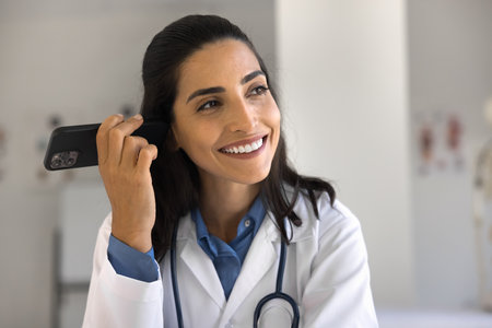 Positive beautiful young doctor woman listening to audio voice message from patient on mobile phone, using digital device for work communication in medicine, healthcareの写真素材