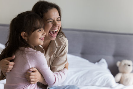 Overjoyed mother hugging laughing little 5s daughter sit on bed in bedroom, look away, having fun in morning at home, enjoy playtime, have playful mood feel happy spend family leisure indoor, close upの写真素材