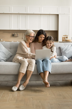 Positive excited grandmother, mom and little tween child using laptop on home couch together,...