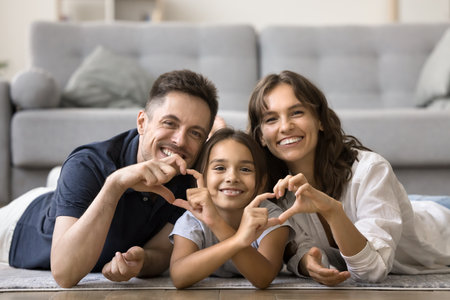 Cheerful attractive mom, dad and sweet daughter kid joining fingers, showing hand heart shapes,...