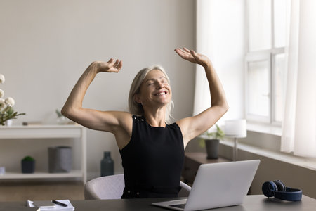 Happy middle aged business woman sitting at desk with eyes closed, rising hands, stretch back after workday finishing, satisfied with good job done, plan weekend, feel successful completion of projectの写真素材