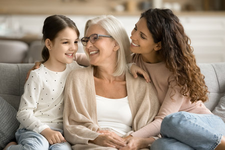 Cheerful grandma, mom and grandkid girl resting on home couch, talking, laughing, having fun, enjoying family leisure, warm close relationship, happy childhood, retirementの写真素材