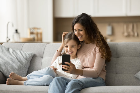 Cheerful pretty black haired mom and positive daughter child talking on video call on smartphone, resting on home couch, using mobile phone for online family communicationの写真素材