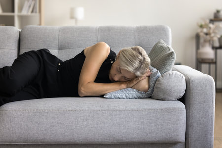 Mature woman resting on sofa take nap in living room. Unhappy middle-aged single female lying down on couch feel stressed, unhappy after break up, fight, going through divorce, coping with depressionの写真素材
