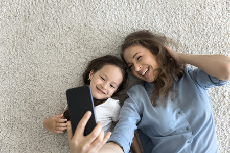 Happy mother and daughter lying on floor looking at cellphone screen, take selfie pictures, having fun using new mobile application, spend free time on internet, make videoconference call, top viewの写真素材