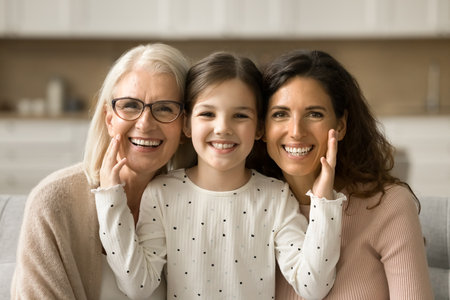 Cheerful cute little girl, mom, grandmother sitting close indoors, hugging, looking at camera with toothy smiles. Happy daughter kid touching cheeks of younger mother and grandma head shot portraitの写真素材