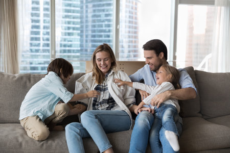 Loving parents play with two siblings at home. Young mother and father enjoy funny playtime with preschoolers son and daughter, laughing, tickling each other, feel overjoyed. Family games and leisureの写真素材