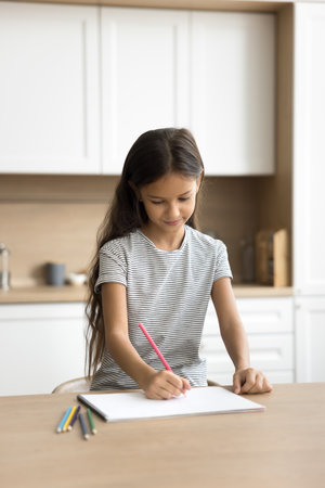 Positive pretty little kid girl drawing in colored pencils at kitchen table, enjoying creativity, artistic hobby, doing art school homework task, scratching doodles in paper albumの写真素材