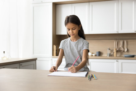 Focused preteen kid girl drawing colorful sketches, doodles, sitting at kitchen table at modern home, doing school homework task, training creative artistic skills, practicing educational activityの写真素材