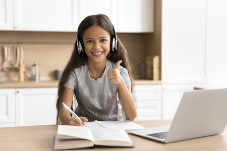 Happy elementary school pupil home headshot portrait. Cheerful beautiful young preteen girl in headphones sitting at laptop, studying on Internet, looking at camera, showing thumb up like gestureの写真素材