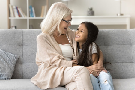 Happy loving blonde grandma and cute preteen granddaughter kid talking and laughing on couch, enjoying funny discussion, affection, trust, family leisure. Grandmother meeting with girl at homeの写真素材