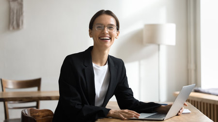 Happy young professional woman workplace head shot portrait. Cheerful businesswoman, project manager in formal suit sitting at laptop, looking at camera with toothy smile, laughingの写真素材