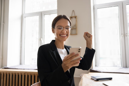 Cheerful successful young business woman holding mobile phone, making winner yes hand gesture, feeling happy, joyful about good news, celebrating success, achievement, win at office workplaceの写真素材