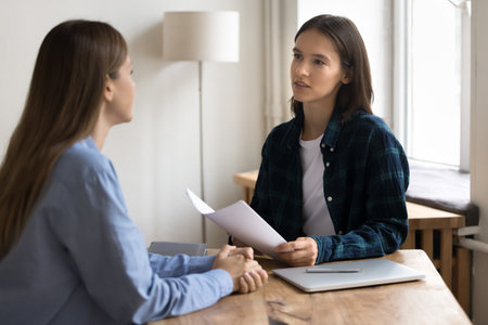 Serious young project manager woman in casual informal clothes talking to female business colleague at work table, using paper documents, holding resume, interviewing job candidate,の写真素材