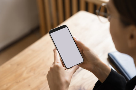 Blank screen of smartphone in young business woman hands. Female employee, manager using mobile phone, holding gadget over workplace office, looking at blank white touch screenの写真素材