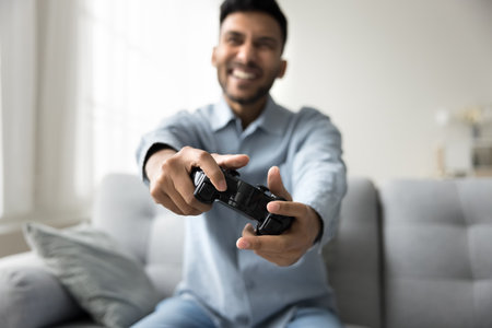 Happy cheerful young Indian male gamer enjoy exciting videogame at home, playing console online video games seated on sofa with gamepad in hands. Hobby, weekend activity, amusements using modern techの写真素材