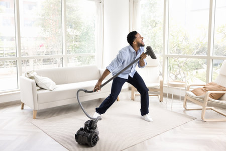 Excited funny young African homeowner man singing at hoover tube like at microphone, cleaning floor at home, enjoying household activities, vacuuming, having funの写真素材