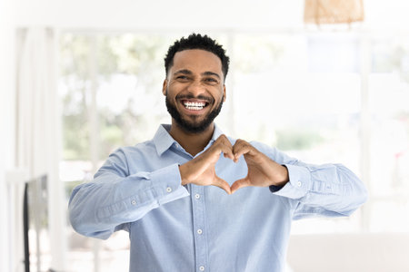 Positive attractive young African man making hand heart gesture, joining fingers at chest, expressing love, affection, support, looking at camera with toothy smile for portraitの写真素材