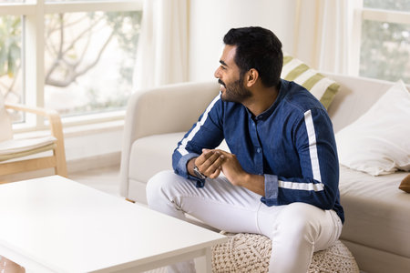 Dreamy relaxed young Indian guy sitting in cozy living room, looking at window away with toothy smile, enjoying home leisure, comfort, relaxing, thinking, dreaming, planning, laughing,の写真素材