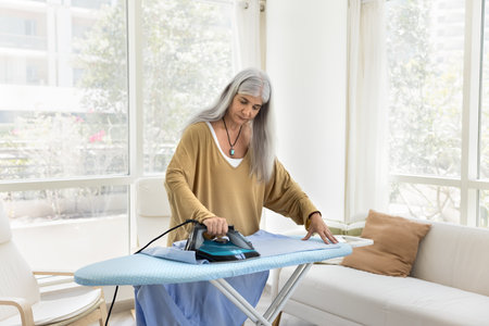 Serious senior Latin maid woman ironing clothing in living room, pressing hot iron to board, using electrical equipment, doing housework, household chores in home apartmentの写真素材