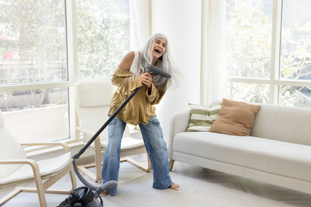 Cheerful excited senior Latin maid woman having fun while cleaning apartment, vacuuming floor in living room, singing at brush of cleaner, dancing to music enjoying funny domestic houseworkの写真素材