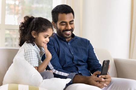 Cheerful loving Indian daddy showing funny video to cute daughter kid at home, talking on family conference call. Father and sweet little girl using smartphone for online communication togetherの写真素材