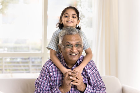 Cheerful pretty little granddaughter girl hugging grandpa from behind. Happy Indian grandfather...
