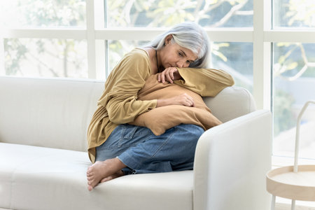 Depressed retired elderly woman sitting on couch at home, keeping closed posture, embracing cushion, feeling bad, lonely, sad, frustrated, looking away with heavy thoughtsの写真素材