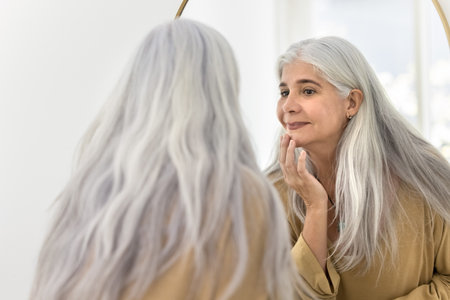 Positive focused elderly Latin woman looking in mirror, touching chin, reviewing skincare procedures results, smiling. Back view and reflection of senior beauty care model with naturally grey hairの写真素材