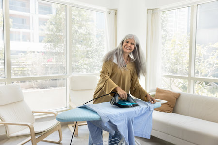 Cheerful positive retired senior Latin woman enjoying household chores at home, ironing fresh dry clothes on board, looking at camera, smiling, doing domestic housework in apartmentの写真素材