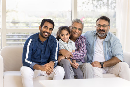 Happy Indian great grandpa holding little kid girl in arms on lap, sitting at mature son and young adult grandson on sofa, looking at camera. Four of family generations posing for home portraitの写真素材