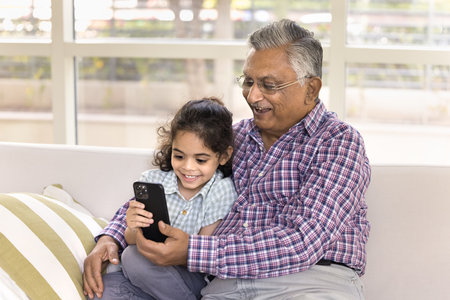 Happy Indian grandpa and cute little granddaughter kid watching funny content on mobile phone, using...